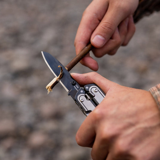 A man using a Leatherman ARC MagnaCut knife blade to take bark off a tree branch.
